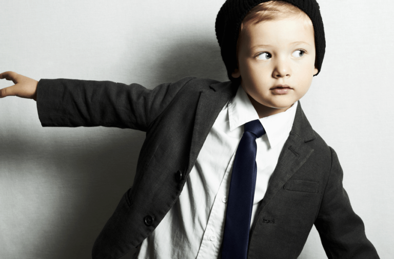 A stylish young boy in a suit and tie, wearing a beanie, poses confidently against a simple background.