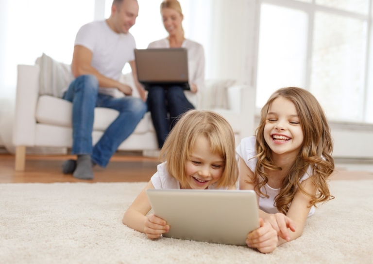 Two smiling girls lie on the floor, using a tablet, while their parents work on laptops in the background.