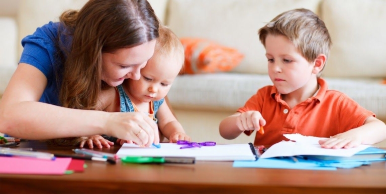 A mother helps her two children with an arts and crafts project, surrounded by colorful papers and markers at a table.