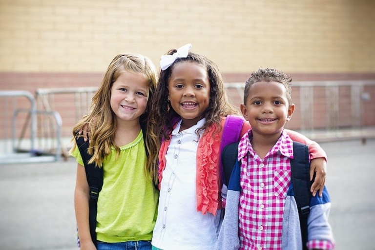 Three smiling children with backpacks, standing together outdoors, showcasing friendship and joy.