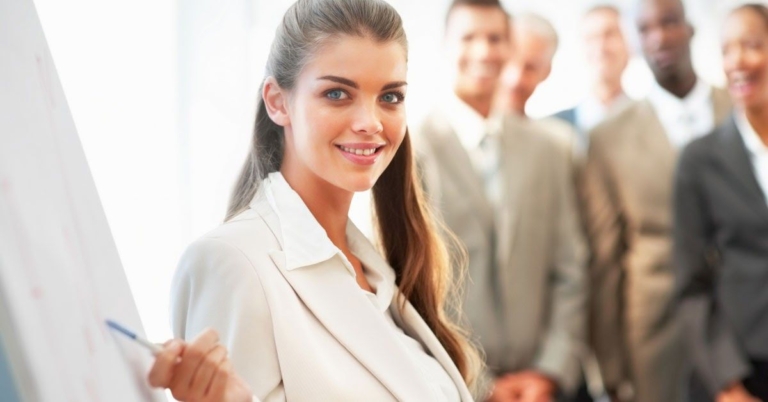 A confident woman in a suit smiles while presenting to a group of professionals in a bright, modern setting.