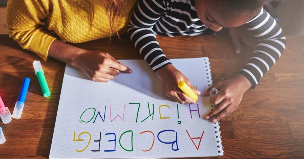 A child and an adult write colorful letters on a large sheet of paper, showcasing creativity and collaboration.