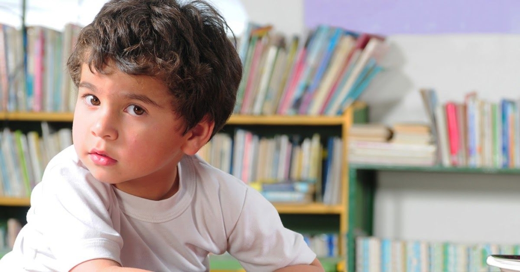 A young boy with curly hair looking thoughtfully at the camera, seated in a library surrounded by books.