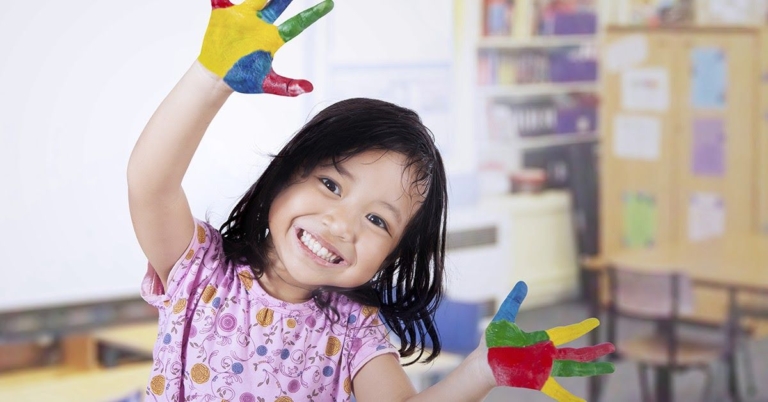 A cheerful girl with painted hands, smiling widely and showing off her colorful artwork in a classroom setting.