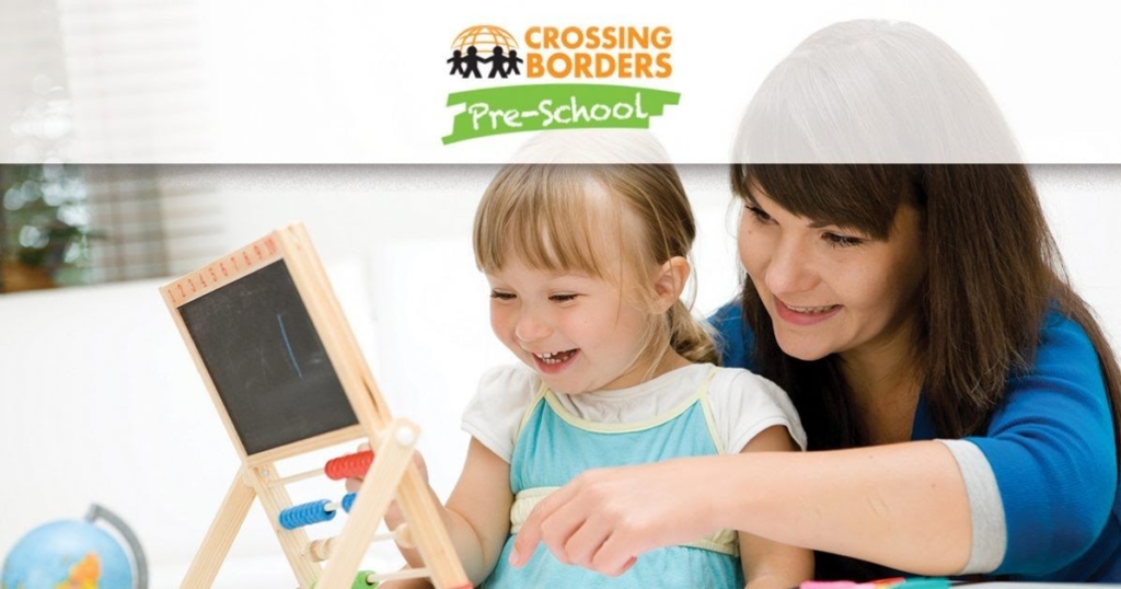 A teacher smiles while helping a young girl with a chalkboard, highlighting joyful learning in a preschool setting.