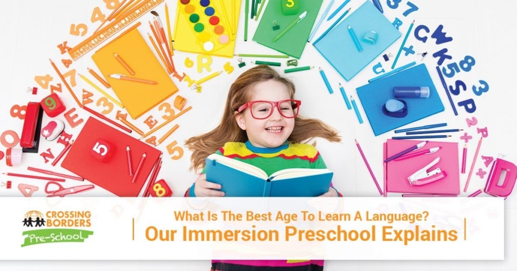 A joyful child in colorful glasses surrounded by school supplies, smiling while reading a book about language learning.