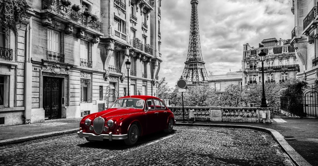 A vintage red car on a cobblestone street, with the Eiffel Tower in the background, set in a black-and-white scene.