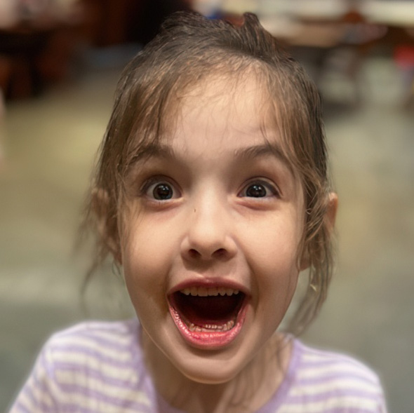 A girl with long hair excitedly opens her mouth in a big smile, displaying pure joy and enthusiasm in her expression.