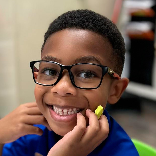 A cheerful boy in glasses smiles widely, resting his chin on his hand, radiating playfulness and joy.