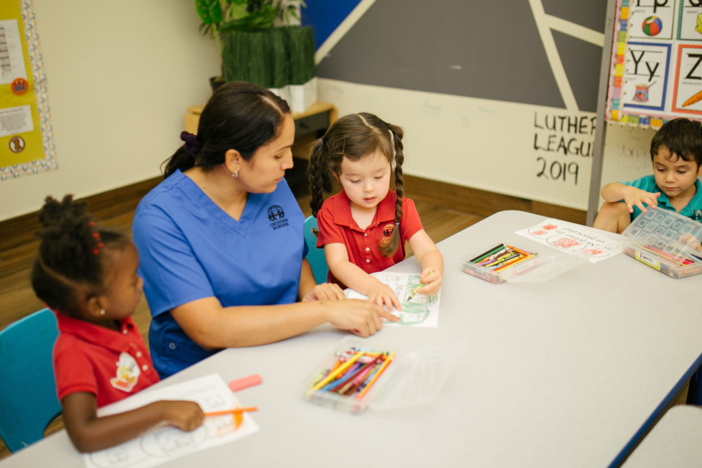 A teacher assists three young children with coloring activities at a table, fostering creativity and learning in a classroom.