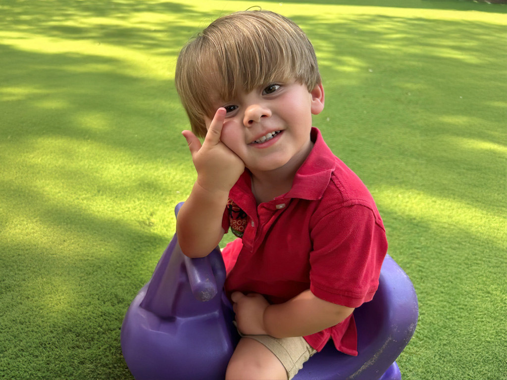 A young boy plays joyfully on a purple toy in a vibrant green area.