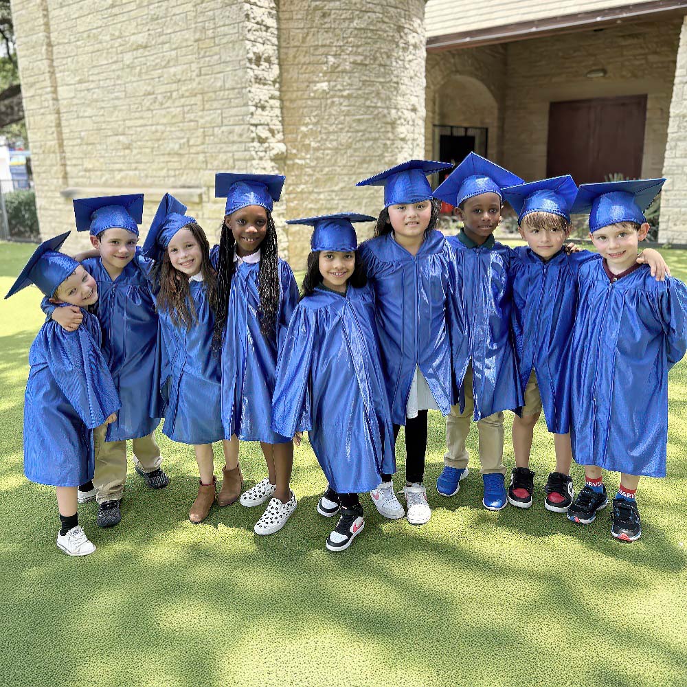 A group of children in blue graduation gowns and caps smiles together, celebrating their achievement outdoors.