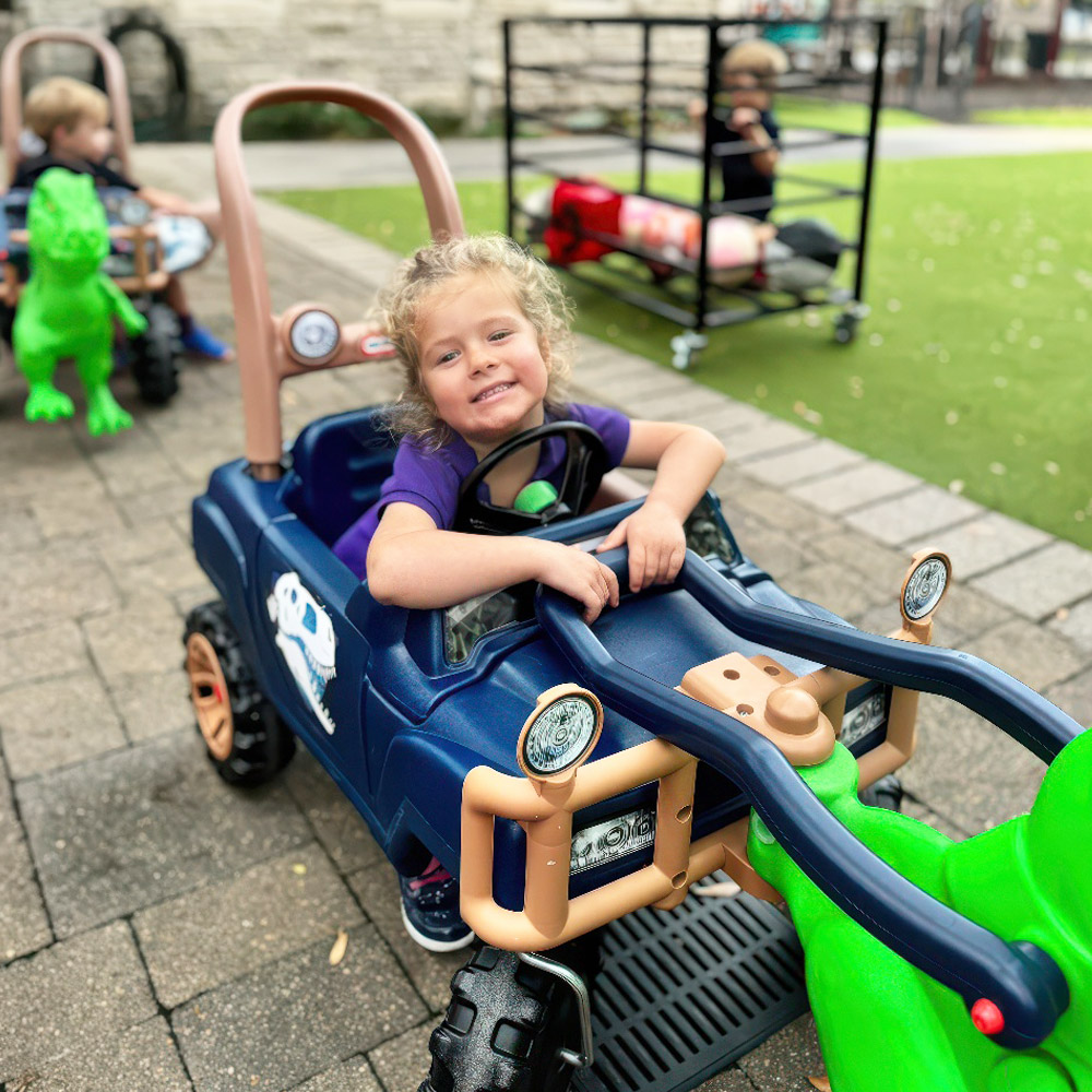 A smiling girl leans on a toy truck, enjoying outdoor playtime and adventure with friends in the background.