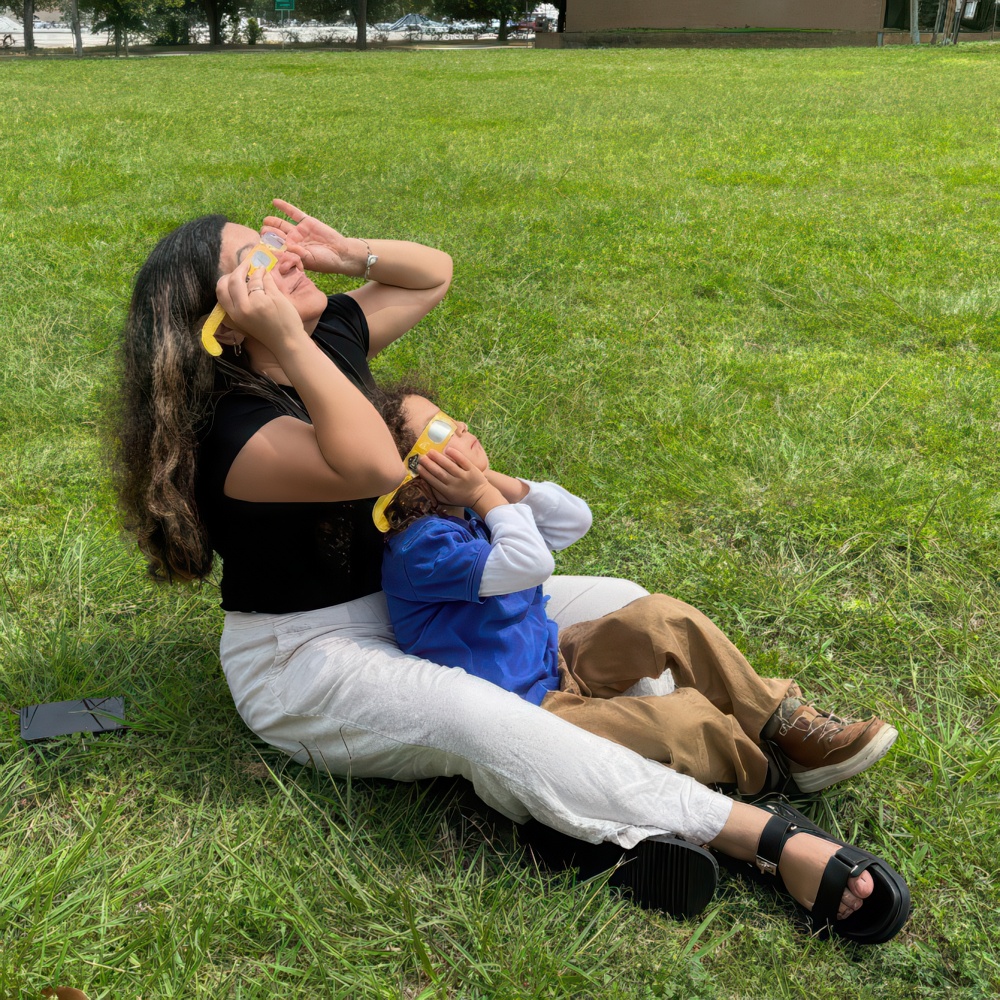 A teacher and a child sit on grass, both holding toy phones to their ears, enjoying a playful moment together.