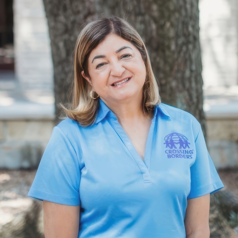 A smiling woman in a light blue shirt stands outdoors, radiating warmth and friendliness against a natural background.