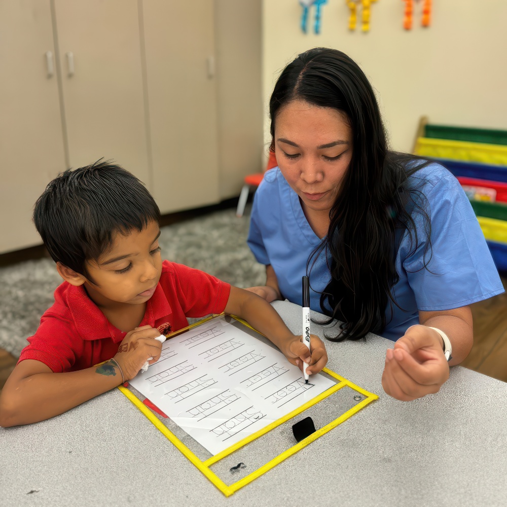 A teacher guides a young boy as they work on a writing activity together at a table, focused and engaged in learning.