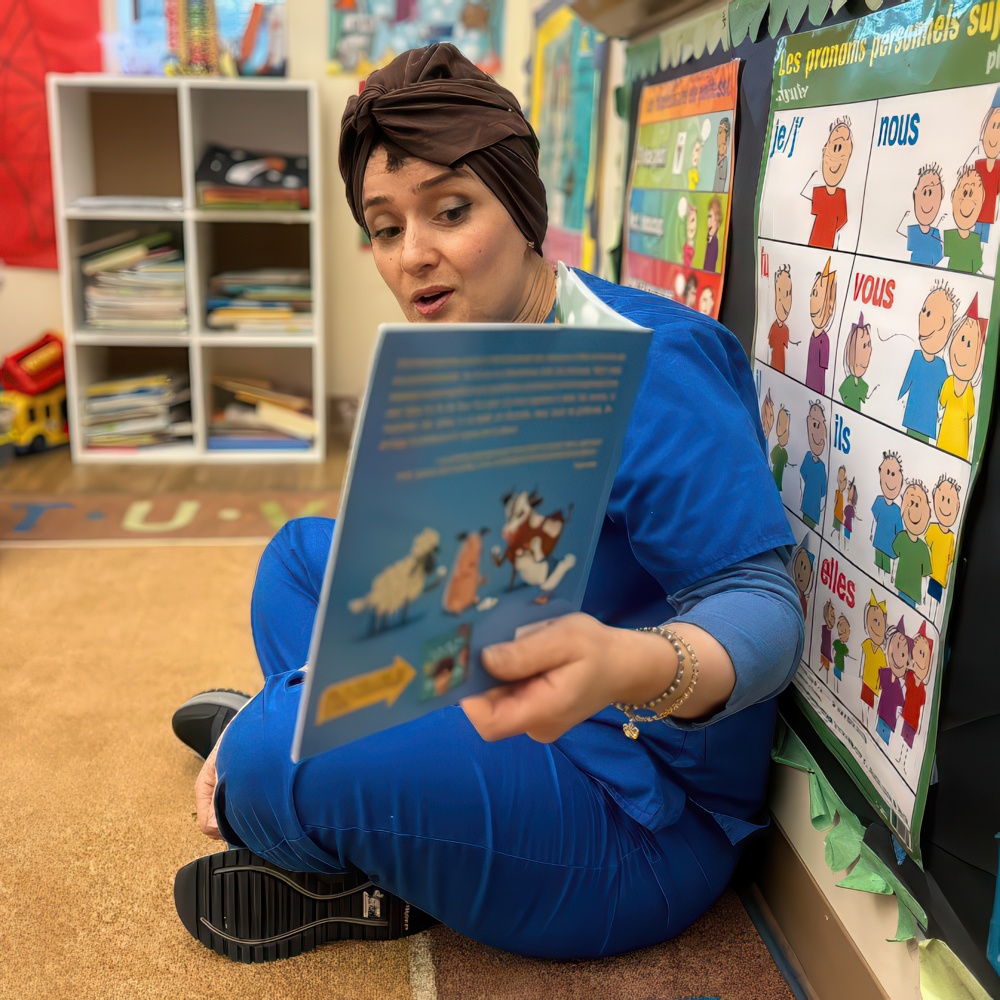 A teacher in blue uniform reads a book while seated on the floor, engaging in a colorful, educational setting.