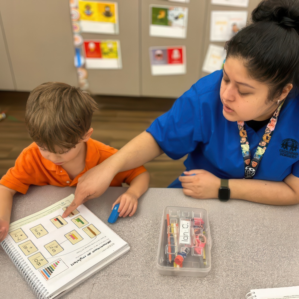 A teacher helps a young boy with a lesson, pointing to illustrations in a book while he follows along, focused on learning.