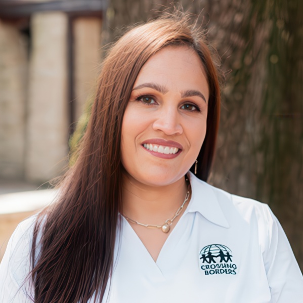 A woman with long, straight hair smiles brightly, wearing a white shirt with a logo, set against a natural background.