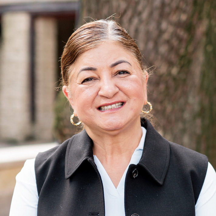 A smiling woman in a black vest poses outdoors, radiating warmth and friendliness against a natural backdrop.