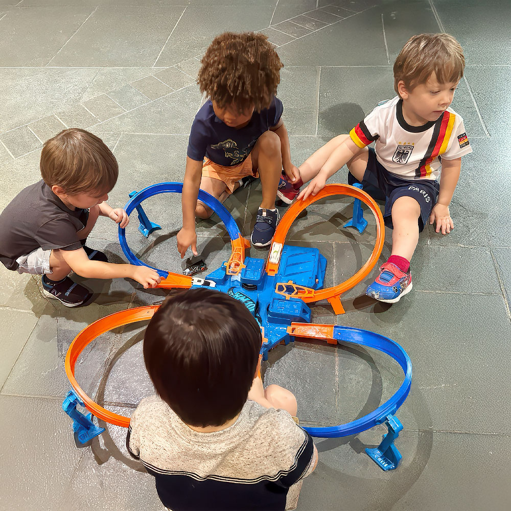 Four children collaborate on a colorful toy race track, engaged in play and teamwork as they set up their cars.