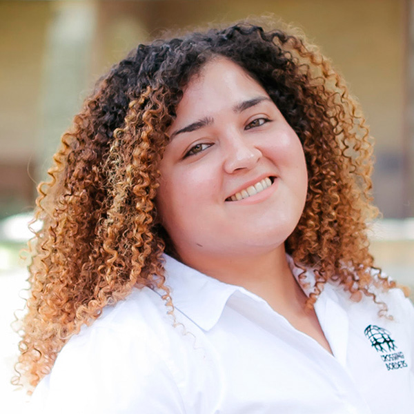 A woman with curly, voluminous hair smiles brightly, wearing a white shirt against a softly blurred backdrop.