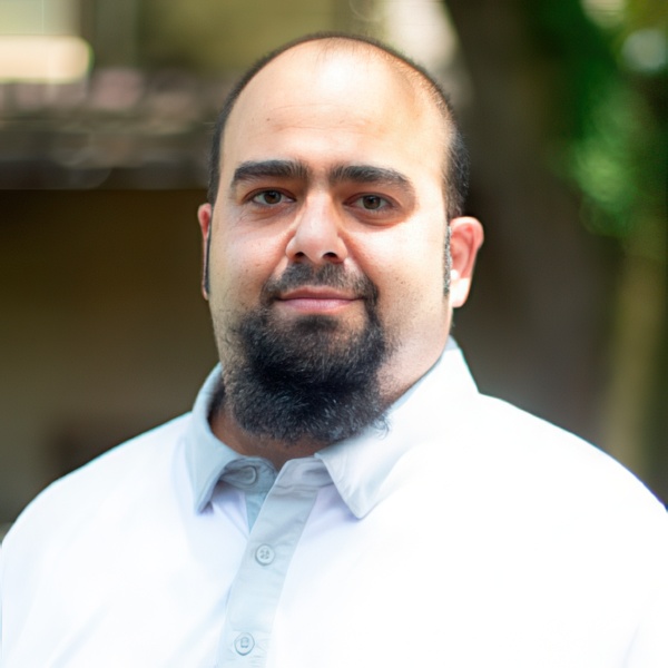 A man with a beard and short hair stands confidently, wearing a light button-up shirt against a blurred outdoor background.