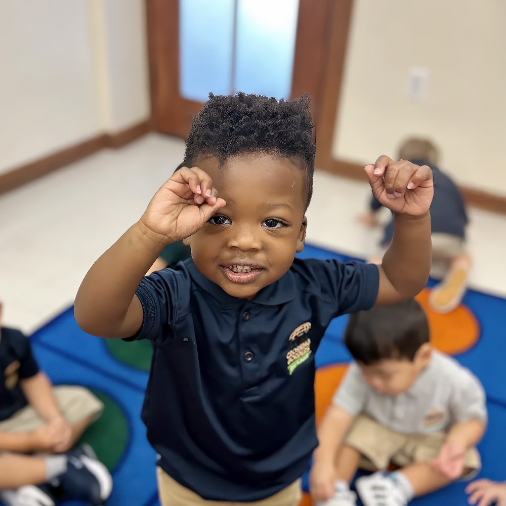 A smiling boy raises his hands in excitement, with other children playing in the background on a colorful mat.