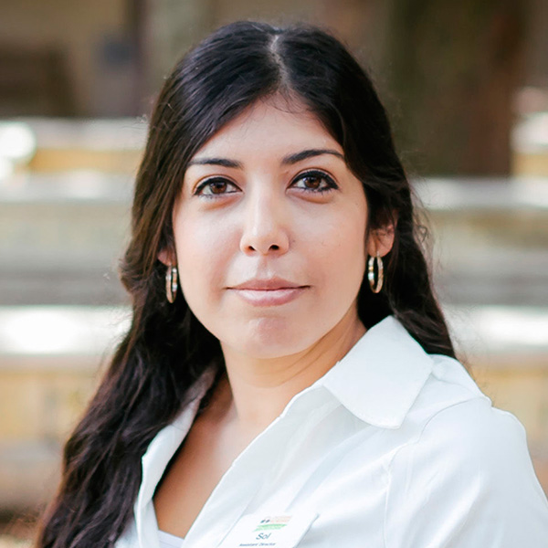 A woman with wavy hair and hoop earrings smiles confidently, wearing a white shirt against a softly blurred background.