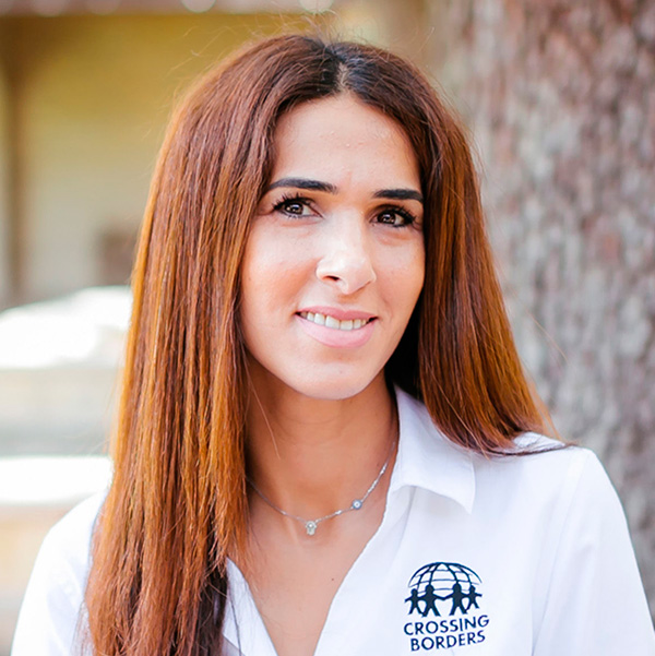 A woman with long, straight hair smiles warmly, wearing a white shirt, and stands against a natural background.