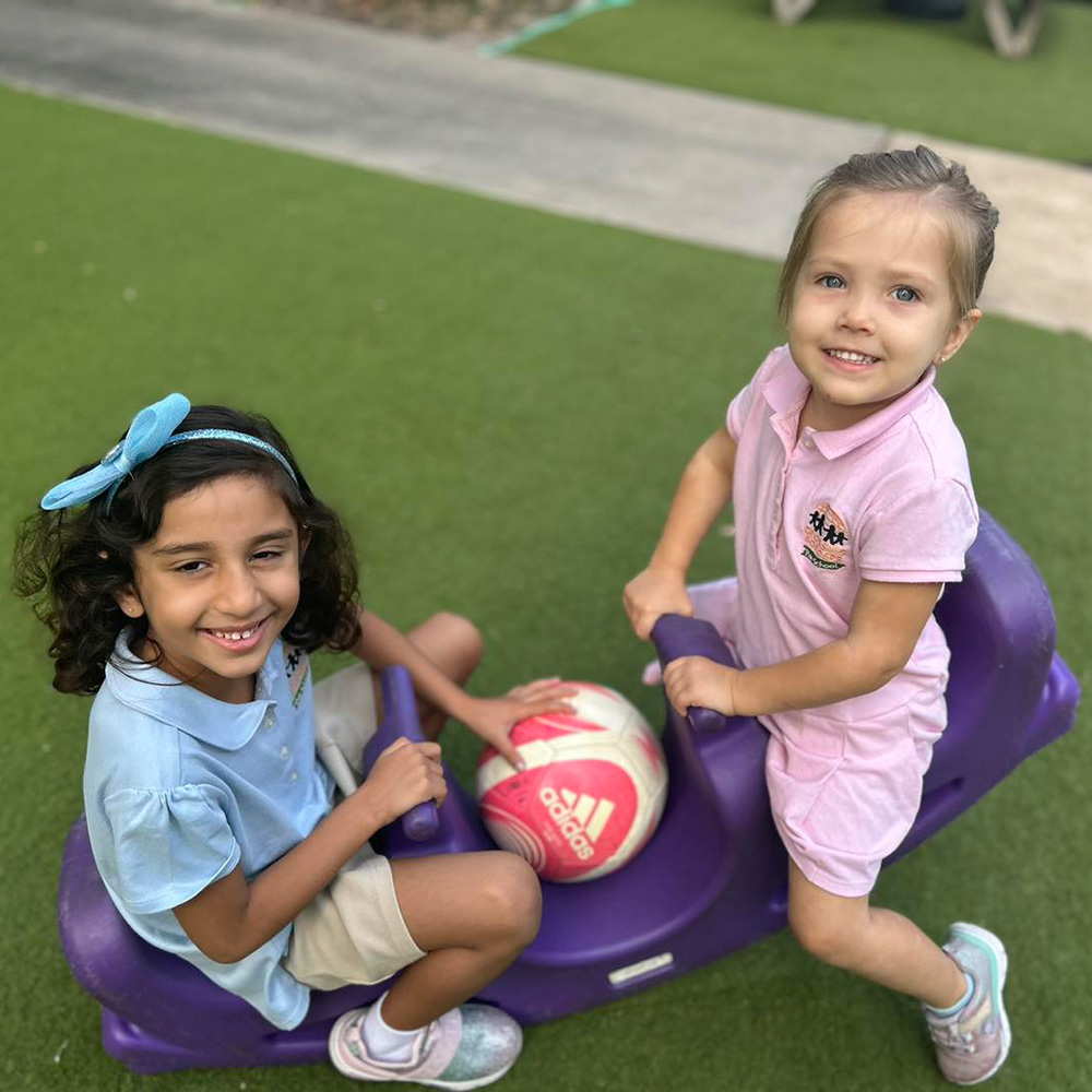 Two girls happily play together outdoors, sharing a soccer ball while seated on a purple ride-on toy.