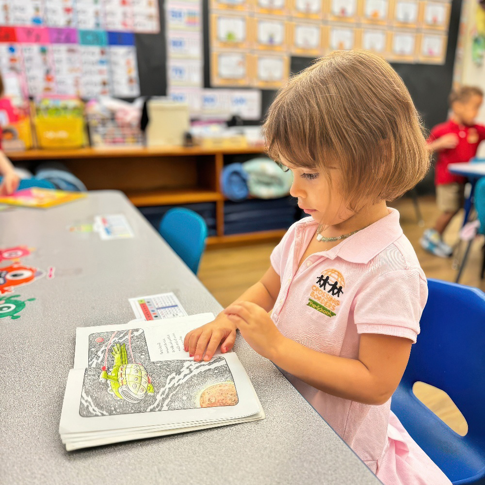 A young girl attentively explores a colorful book at a table, fully engaged in her reading experience.