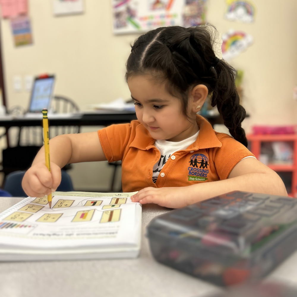 A girl in an orange shirt concentrates as she trace in a workbook, holding a pencil and sitting at a table.