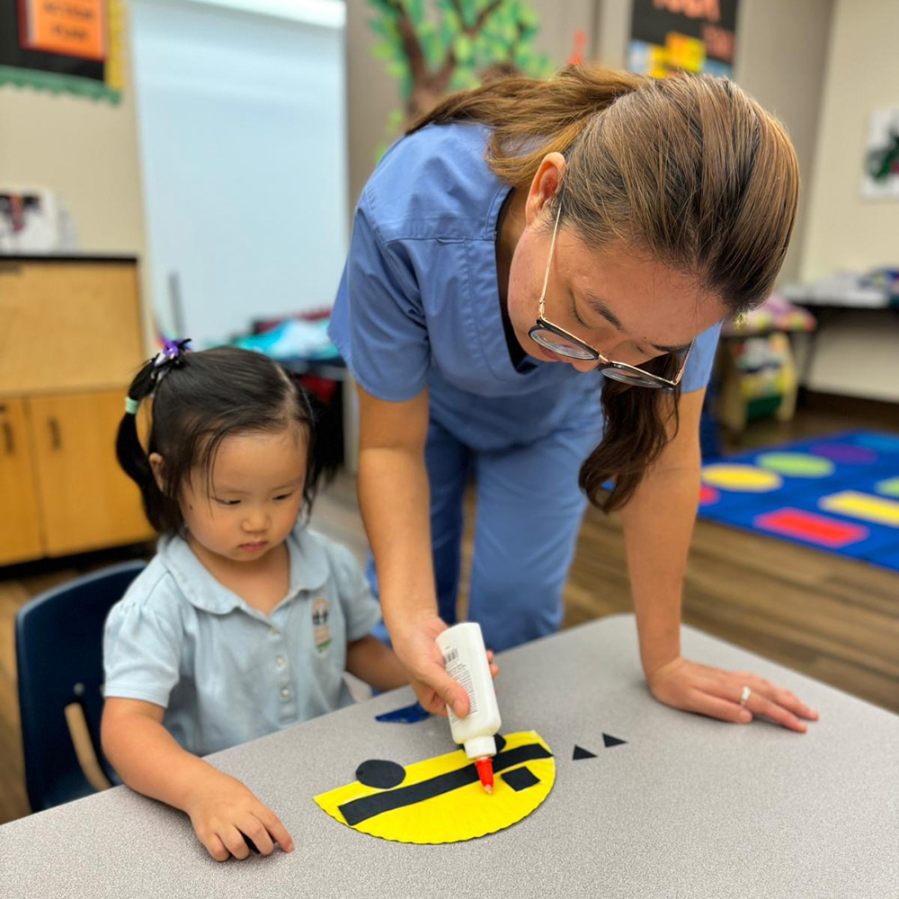 A Teacher shows a child how to use glue for a craft project, both focused on the colorful materials in front of them.