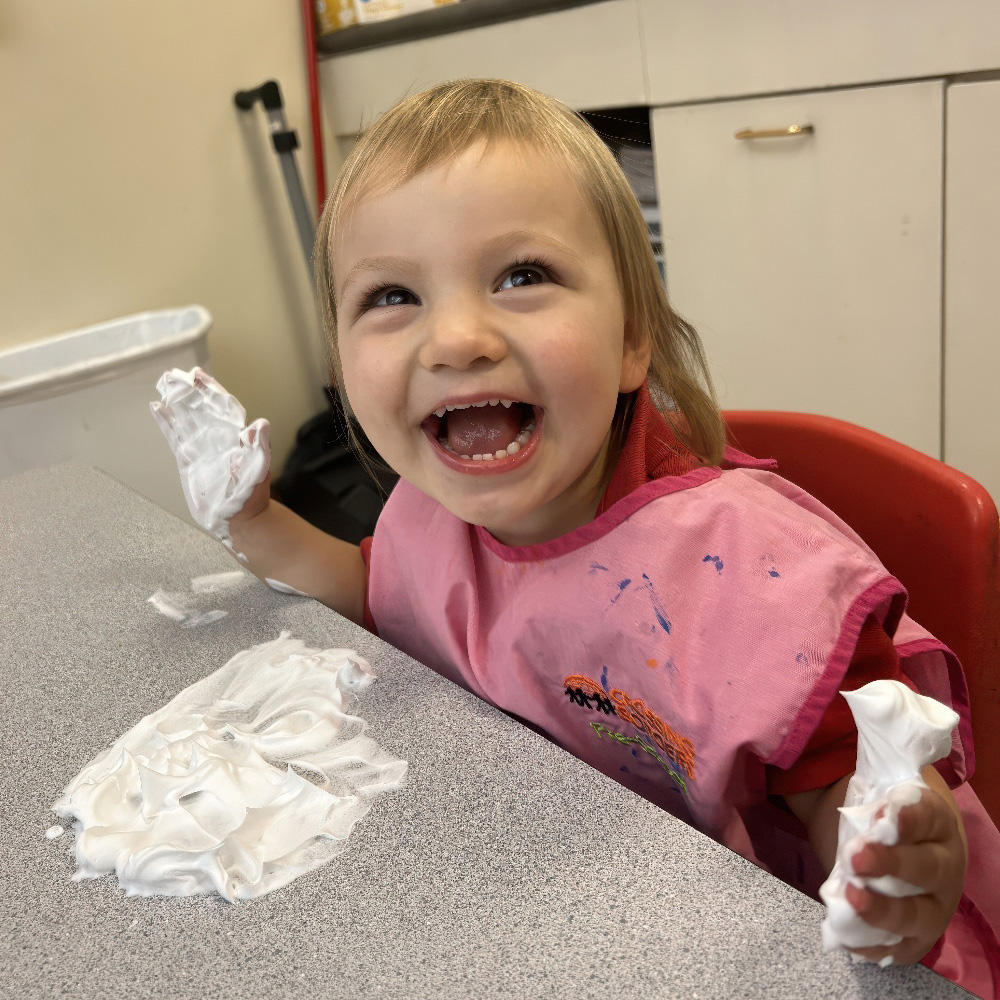 A joyful child in a pink smock plays with shaving cream, smiling widely while creating a fun, messy art experience.