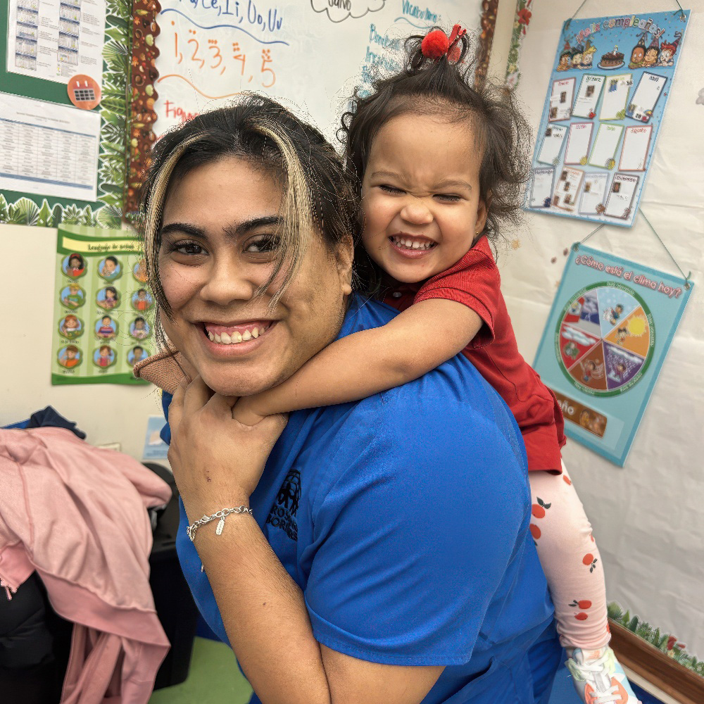 A smiling teacher in blue carries a joyful child on her back, both radiating happiness in a vibrant classroom setting.