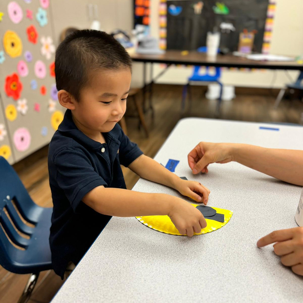 A young boy focuses on a craft activity, placing pieces on yellow paper with guidance from a teacher.