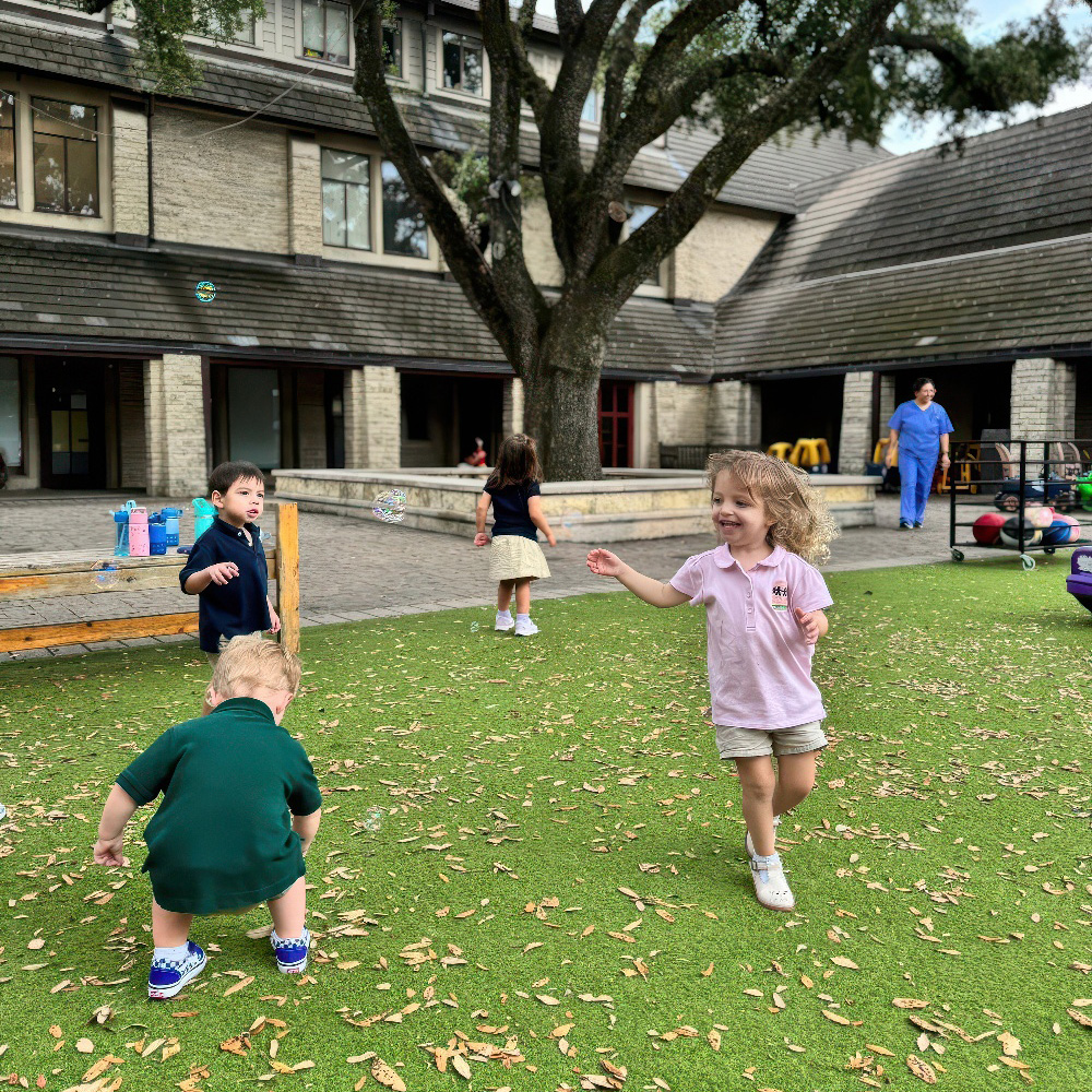 Children play joyfully on a green lawn, running and exploring while a teacher supervises in the background.