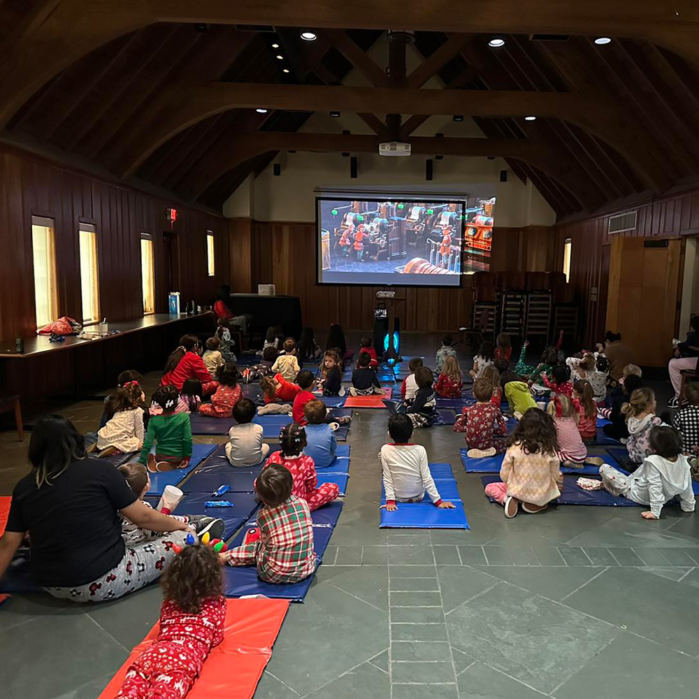 A group of children in pajamas sits on mats, watching a movie on a big screen in a cozy indoor setting.