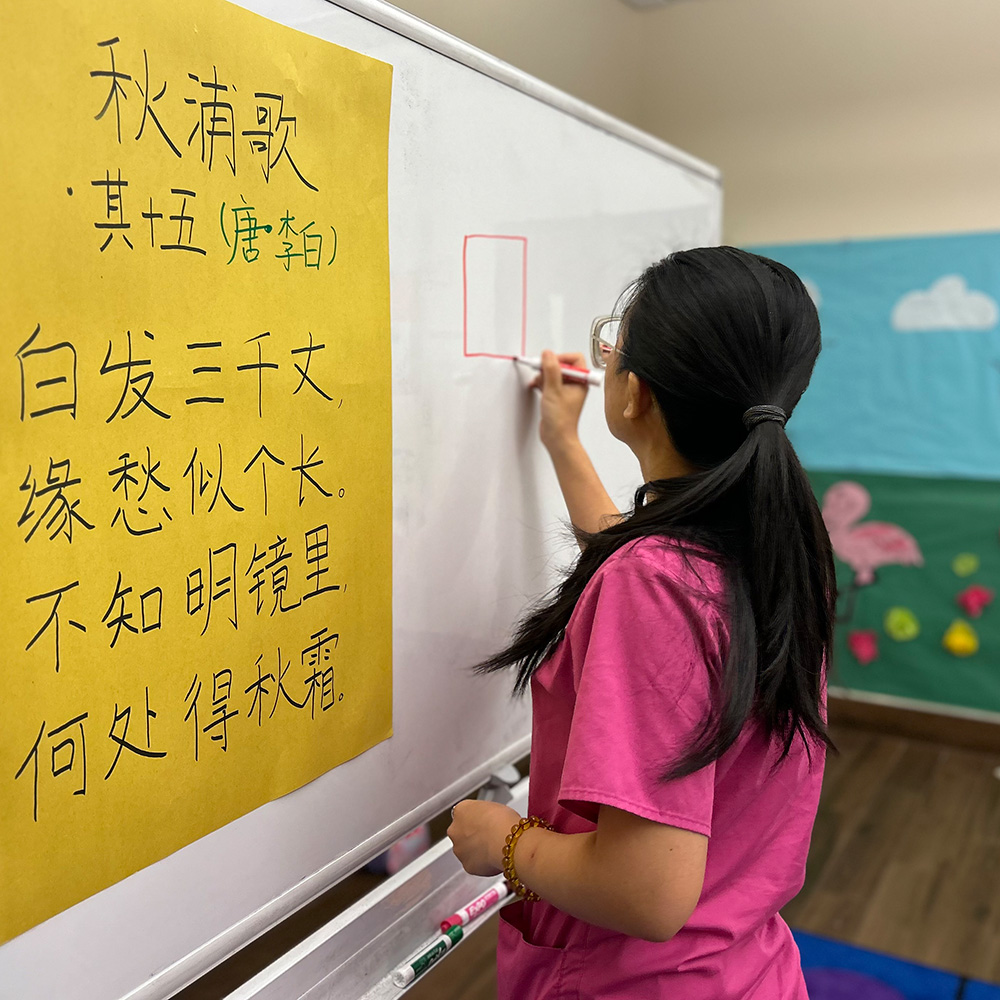 A teacher in a pink shirt draws on a whiteboard, with Chinese text nearby, focused on a creative project.