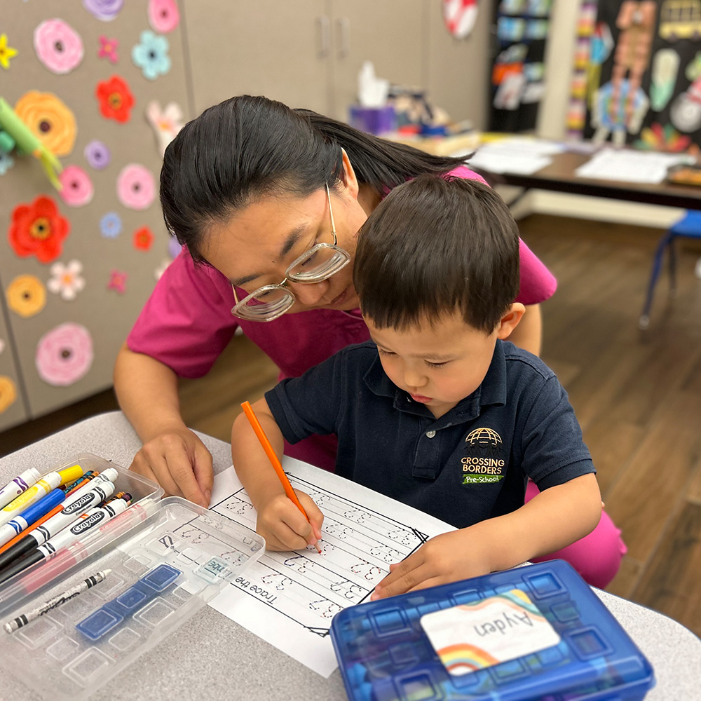 A teacher helps a young boy with a tracing numbers activity, both focused and engaged in their creative task together.