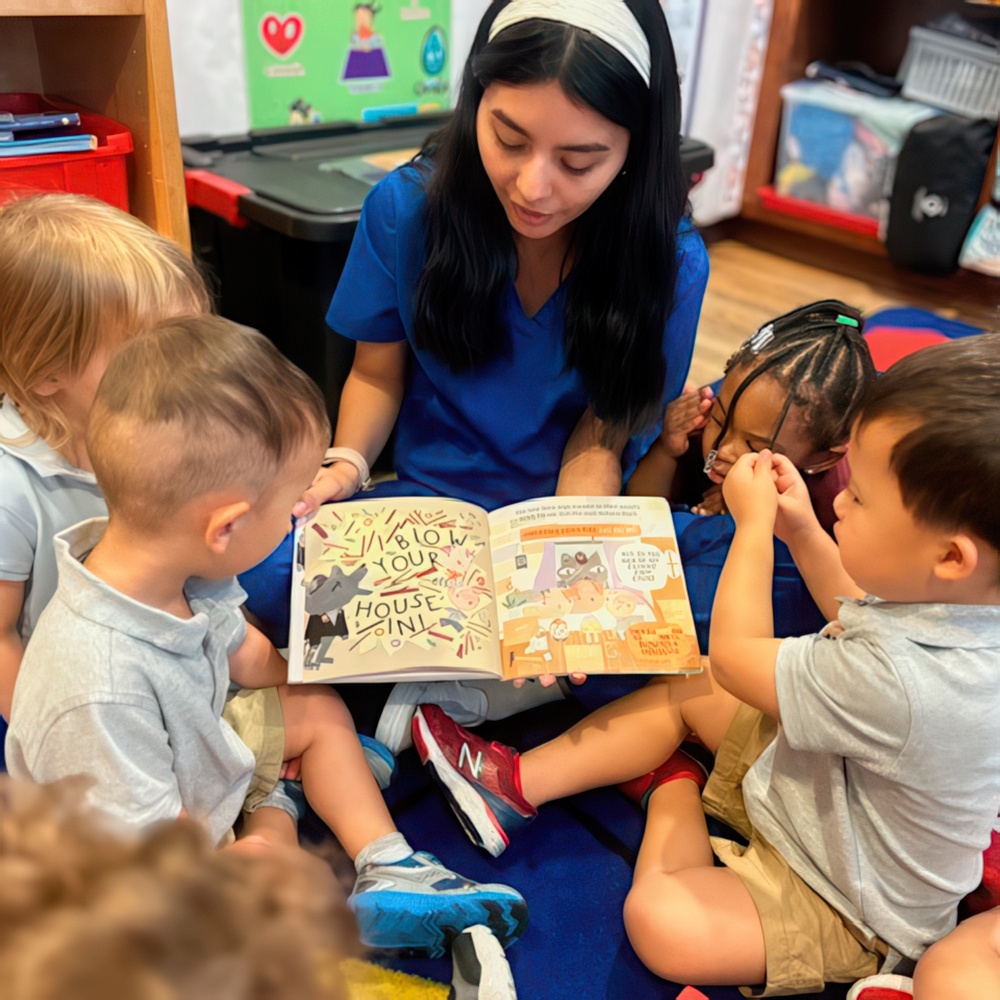 A teacher in blue reads a book to engaged children sitting around her, all focused on the colorful illustrations.