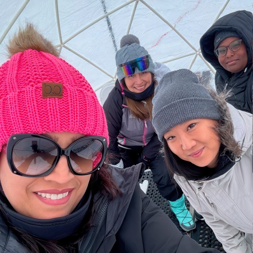 A group of four teacher, bundled in winter gear, smile together inside a cozy igloo, enjoying their snowy adventure.