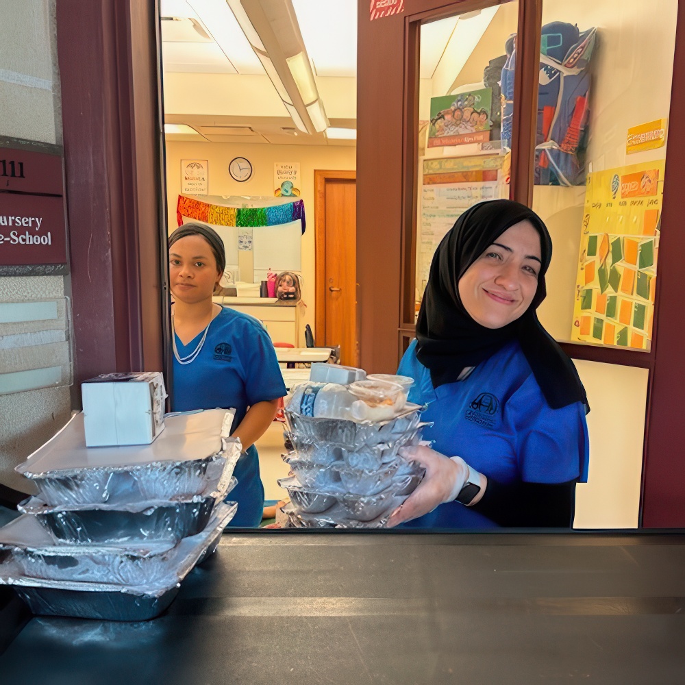 Teachers in blue holds trays of food, smiling as she works in a school kitchen, while another teacher assists behind her.