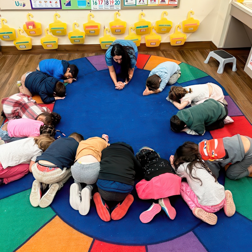 A circle of children with their heads down, engaged in a quiet activity, guided by a teacher in a colorful classroom.