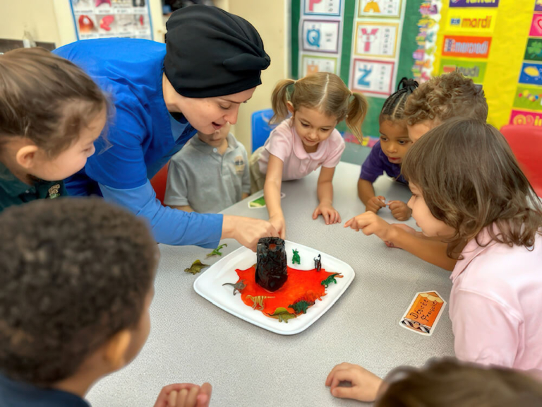 A teacher in blue engages kids around a table, pointing at a colorful project with miniature figures.