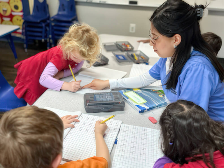 A teacher in blue guides children as they work on grid papers at a classroom table, focused on their tasks.