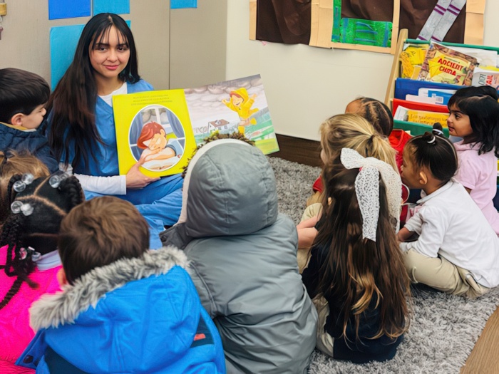 A group of children gathers around their teacher reading a storybook in a cozy classroom setting.