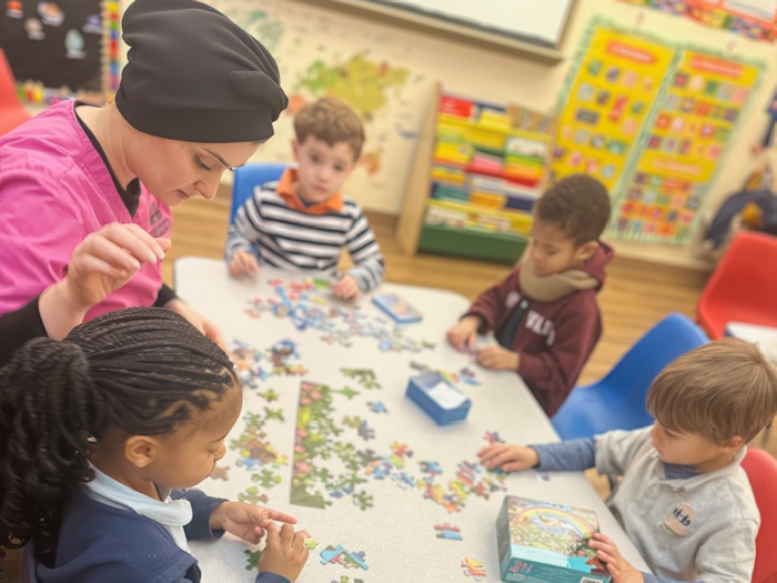 A teacher assists children at a table as they work together on a colorful puzzle in a bright classroom setting.