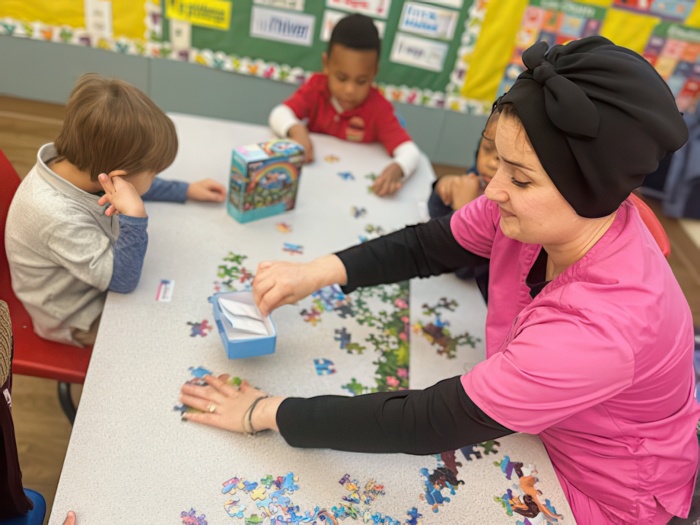 A teacher helps children with a puzzle on a table, while one child observes thoughtfully in a vibrant classroom.